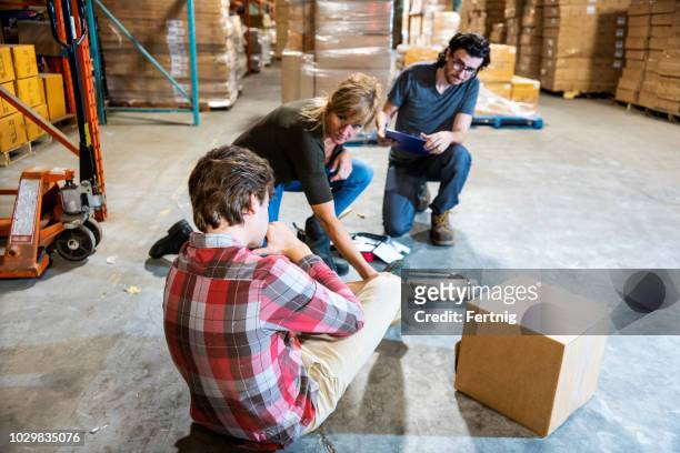 a warehouse worker fallen after tripping on some debris.  he is clutching his ankle in pain as two supervisors assists him. - safety kit imagens e fotografias de stock
