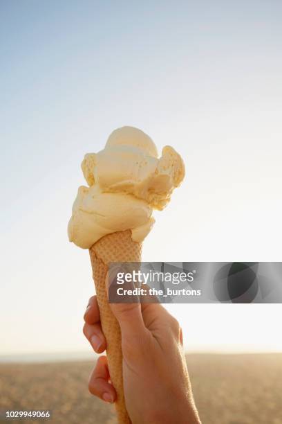 close-up of hand holding ice cream cone with vanilla ice cream at the beach at sunset - frau eistüte stock-fotos und bilder
