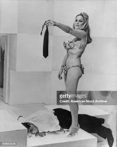Actress Ursula Andress wearing a bikini and high heels while her outstretched hand holds a tie, a man lies beneath her, circa 1965.