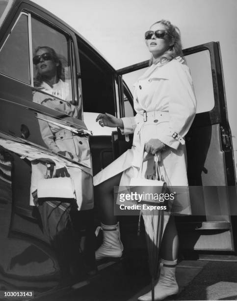 Actress Ursula Andress wearing sunglasses, a white raincoat and boots, as she prepares to enter a car upon her arrival at London Airport, London,...