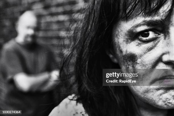 abused and bruised woman with battering partner in background - wife battering stock pictures, royalty-free photos & images