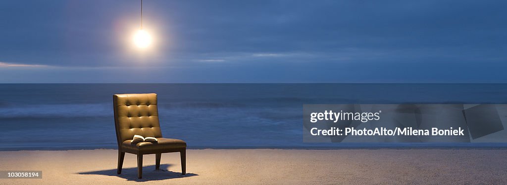 Light bulb illuminated over chair with open book on beach at night