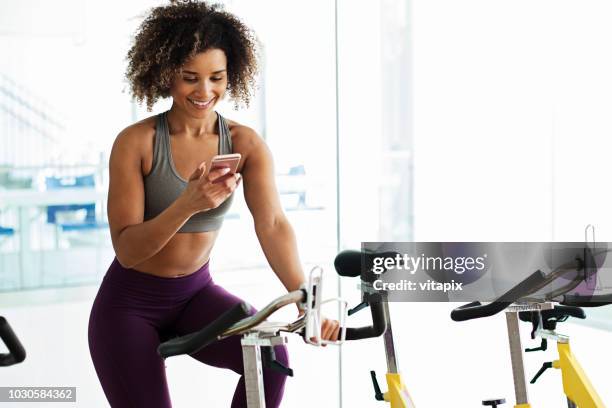 woman on a stationary bike at the gym using her mobile phone - peloton app stock pictures, royalty-free photos & images