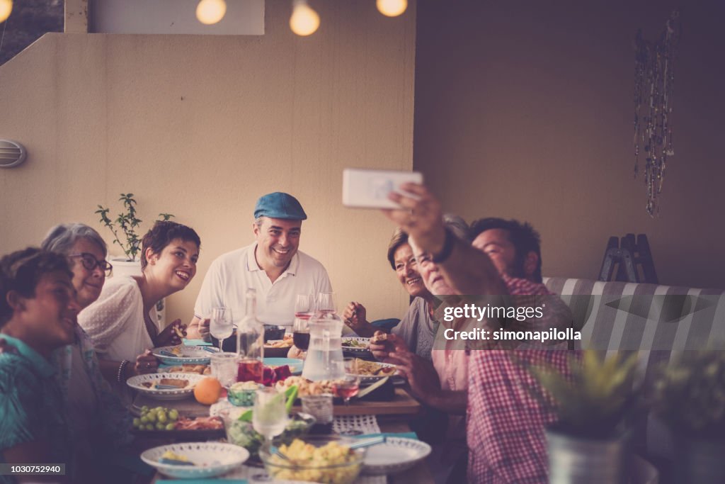 Happy group of people in family friendship all together celebrating and having an outdoor nice dinner at home in the terrace. a man take selfie for all and everybody look at the phone smiling. cheerful people different ages