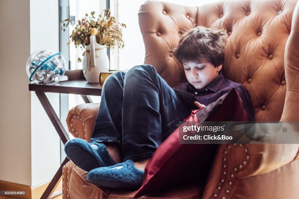 Nine years old boy with tablet in an armchair indoors (home interior)