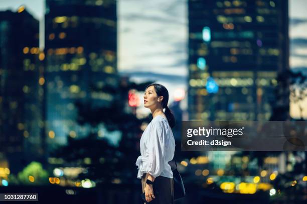 confident young asian woman standing against illuminated city buildings and thinking of her career path - cielo variabile foto e immagini stock