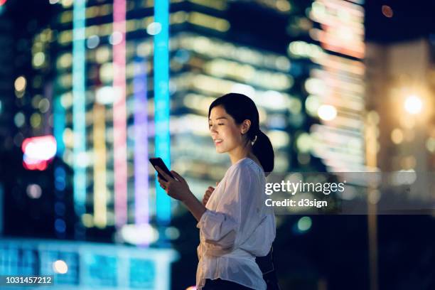 smiling woman checking mobile phone against glowing city skyscrapers at night - central asia stock-fotos und bilder
