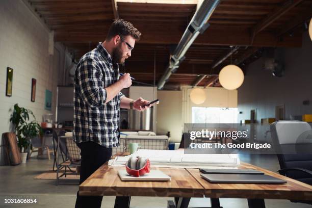 architect standing at desk looking at mobile phone - 2018 blueprint stock-fotos und bilder