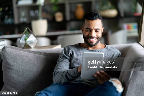 fröhlicher lateinamerikanischen mensch entspannend auf couch beim betrachten social-media auf tablet lächelnd - e reader stock-fotos und bilder