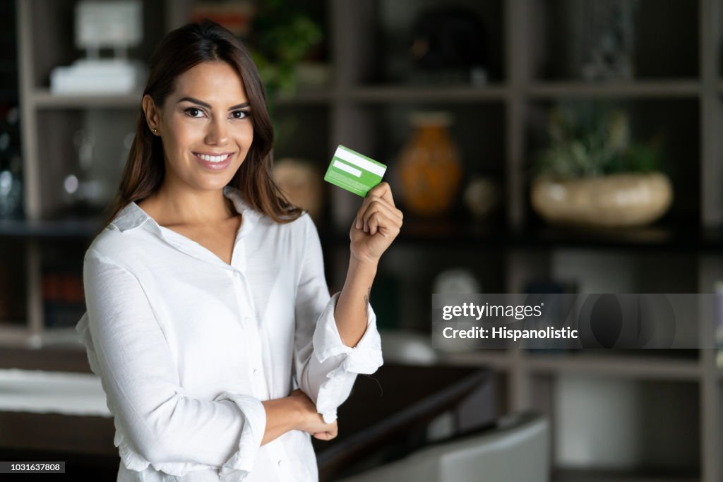 Linda mulher adulta em casa segurando um cartão de crédito e olhando para a câmera muito feliz