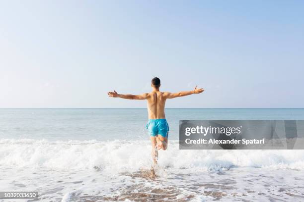 rear view of a young happy man with arms outstretched running towards the sea on a sunny day - swimsuit stock-fotos und bilder