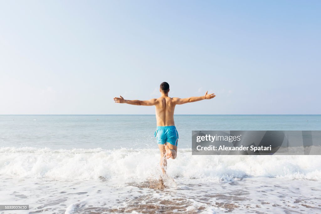 Rear view of a young happy man with arms outstretched running towards the sea on a sunny day