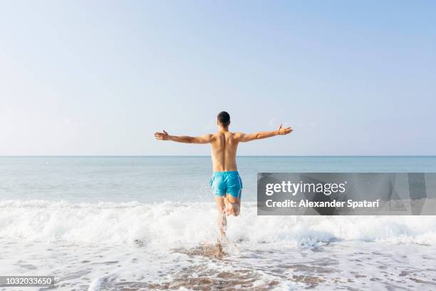 rear view of a young happy man with arms outstretched running towards the sea on a sunny day - men swimwear stock pictures, royalty-free photos & images