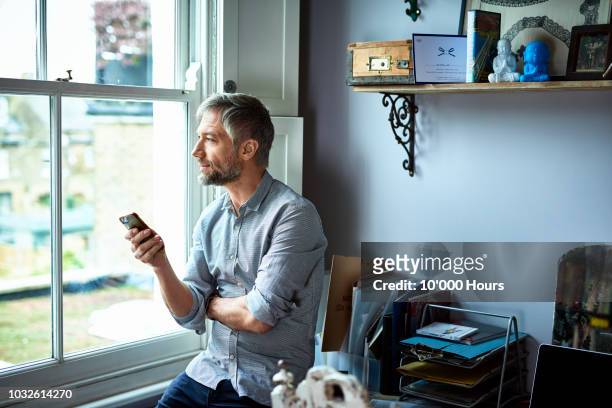 mature businessman using phone in home office looking through window - contemplation stock pictures, royalty-free photos & images