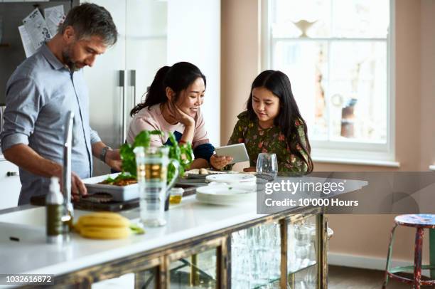 mother and daughter looking at tablet while father prepares home made meal - asian man cooking stockfoto's en -beelden