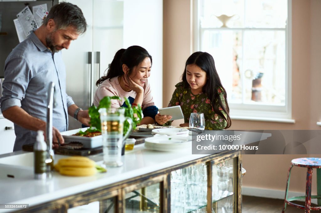 Mother and daughter looking at tablet while father prepares home made meal