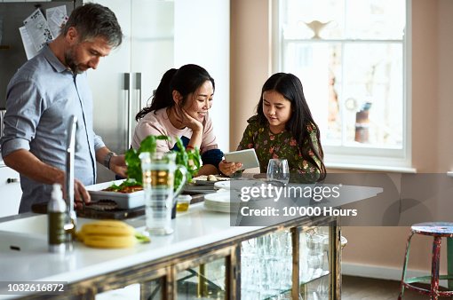 Mother and daughter looking at tablet while father prepares home made meal Mother and daughter looking at tablet while father prepares home made meal
