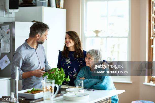 woman with arm around mature woman looking towards man in kitchen - sogra imagens e fotografias de stock