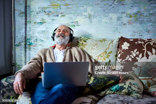 portrait of mature man on sofa smiling and wearing headphones - mature men stock pictures, royalty-free photos & images
