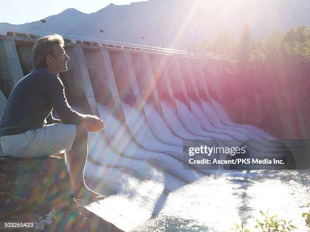 man looks across hydroelectric dam, from rock seat - hydroelectric power stock pictures, royalty-free photos & images