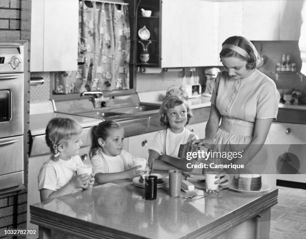 1950s MOTHER THREE DAUGHTERS STANDING AROUND KITCHEN TABLE MAKING LUNCH PEANUT BUTTER JELLY SANDWICHES MOTHER POURING GLASS MILK