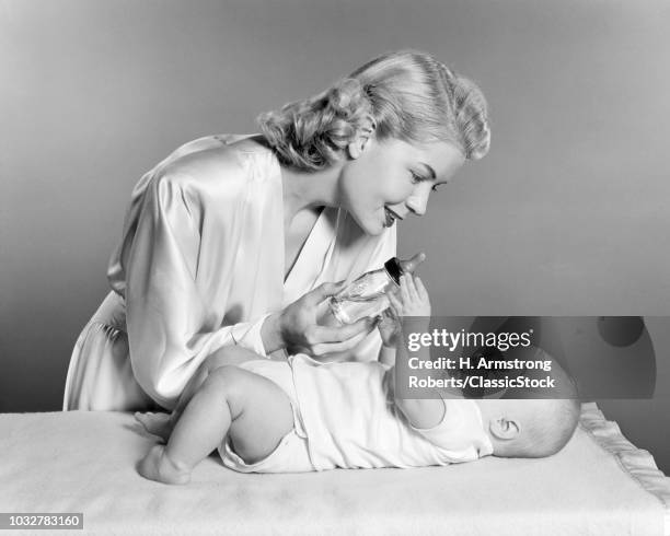 1950s SMILING BLOND WOMAN MOTHER WEARING DRESSING GOWN GIVING BABY BOTTLE FEEDING TO INFANT GIRL DAUGHTER