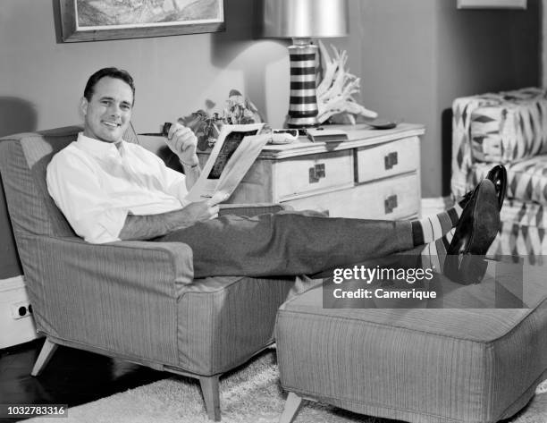 1950s RELAXED MAN SITTING IN EASY CHAIR FEET UP SMOKING PIPE READING MAGAZINE LOOKING AT CAMERA
