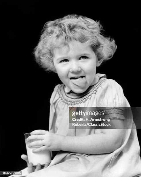 1920s 1930s SMILING LITTLE GIRL CURLY SHORT HAIR HOLDING GLASS OF MILK LOOKING AT CAMERA LICKING HER LIPS WITH TONGUE DELICIOUS