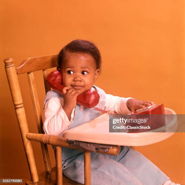 1970s AFRICAN AMERICAN BOY PLAYING WITH TOY TELEPHONE SITTING IN HIGHCHAIR