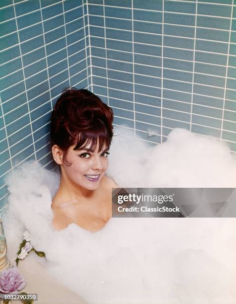 1960s 1970s SMILING WOMAN SITTING IN A BATHROOM TUB OF SOAP BUBBLES AND WATER TAKING A BATH RELAXING INDOOR LOOKING AT CAMERA