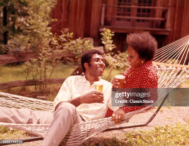 1970s AFRICAN AMERICAN COUPLE MAN WOMAN RECLINING IN HAMMOCK DRINKING BEER OUTSIDE