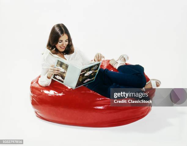 1960s 1970s WOMAN SMILING READING BOOK RECLINING IN A RED BEANBAG CHAIR