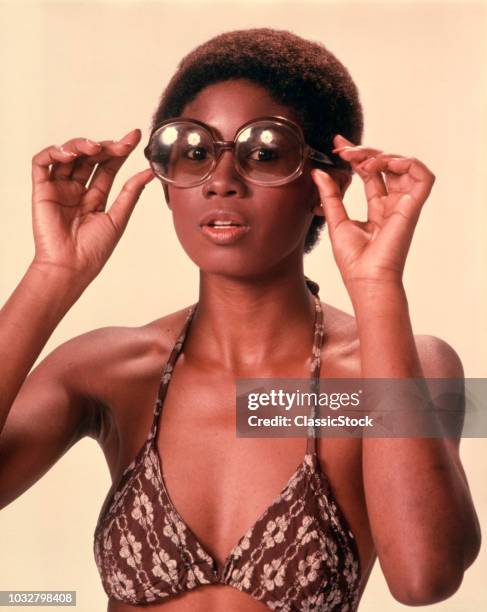 1970s AFRICAN AMERICAN WOMAN WEARING BIKINI BATHING SUIT TOP PUTTING ON LARGE LENS SUNGLASSES LOOKING AT CAMERA