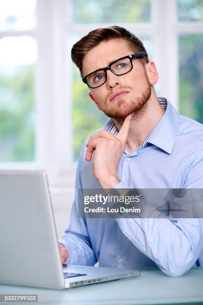 man in a blue shirt, glasses, beard, serious, window out of focus in the background, sitting, in front of a computer laptop; looking up, finger under the chin, looking up. - man looking up beard chin stock-fotos und bilder