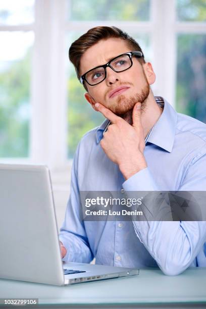 man in a blue shirt, glasses, beard, serious, window out of focus in the background, sitting, in front of a computer laptop; looking up, 2 fingers on the chin, looking up. - man looking up beard chin stock-fotos und bilder