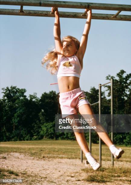 1970s GIRL HANGING FROM MONKEY BARS IN PLAYGROUND WEARING PINK HALTER TOP AND SHORTS