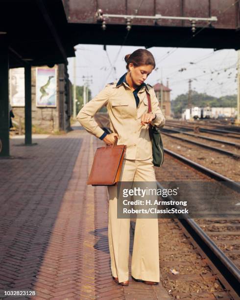 1970s IMPATIENT WOMAN WAITING FOR LATE TRAIN ON PLATFORM WEARING PANTS SUIT HOLDING BRIEFCASE LOOKING AT WRISTWATCH