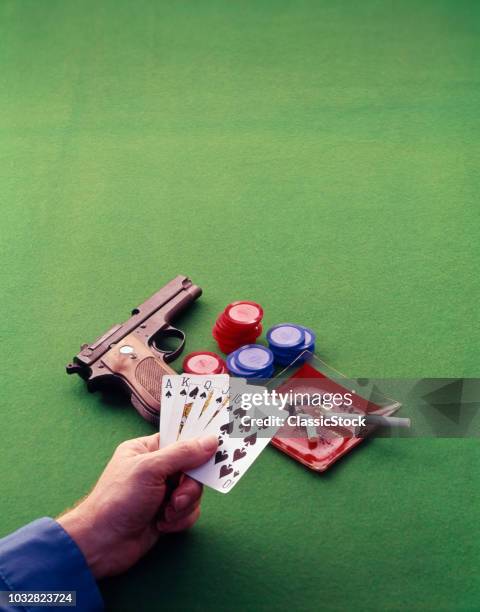 1970s HAND HOLDING ROYAL STRAIGHT FLUSH POKER HAND OVER GREEN FELT CARD TABLE CIGARETTE ASH TRAY CHIPS AND AUTOMATIC PISTOL
