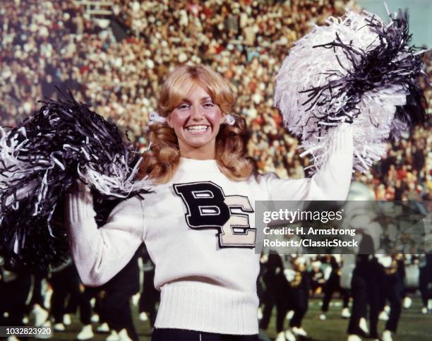 1960s 1970s SMILING BLONDE CHEERLEADER WOMAN BLACK & WHITE SWEATER UNIFORM POMPOMS AT FOOTBALL GAME LOOKING AT CAMERA