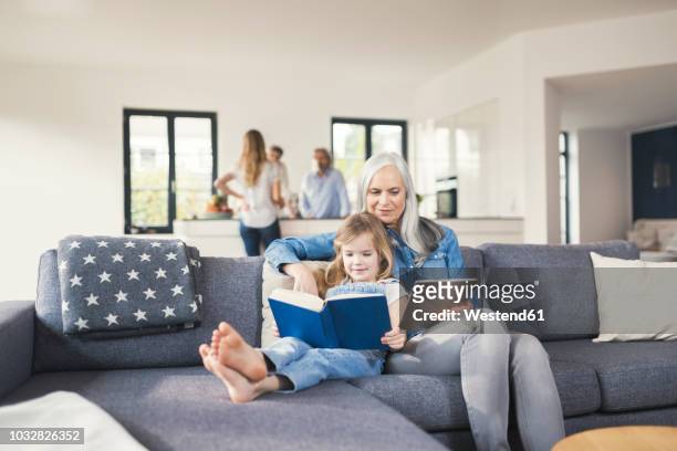 grandmother and granddaughter sitting on couch, reading together book and tablet pc - grandmas living room photos et images de collection