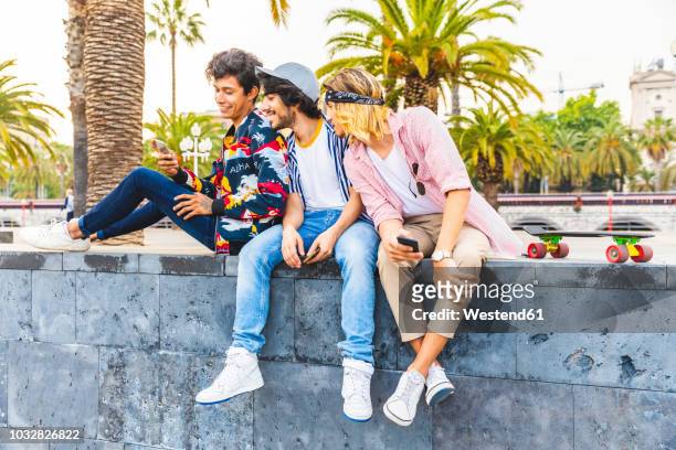 three friends sitting on a wall looking at cell phone - menswear stock pictures, royalty-free photos & images