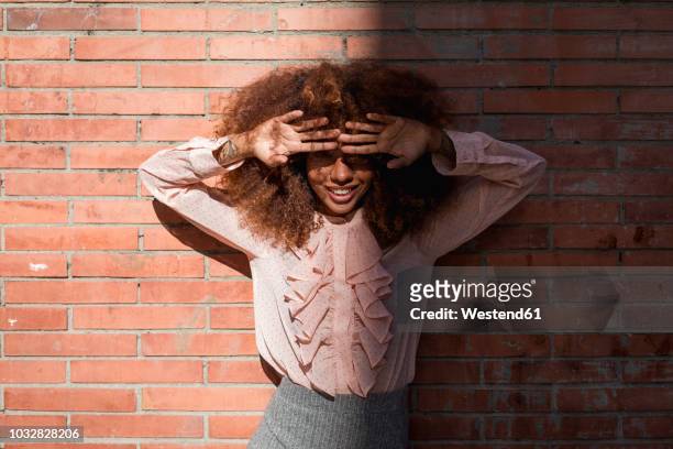 portrait of smiling beautiful young woman with afro hairdoat brick wall in sunshine - afro hairstyle stock-fotos und bilder