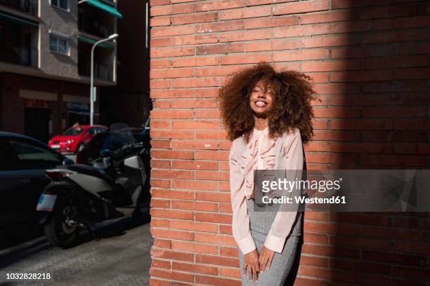 portrait of smiling young woman with afro hairdo leaning against brick wall in the city - afro frisur stock-fotos und bilder