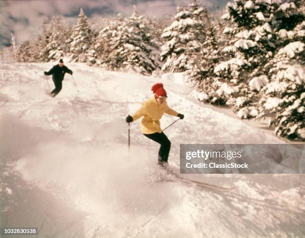 1960s 1970s TWO PEOPLE MAN WOMAN SKIING DOWN SNOWY SLOPE PINE TREES WOMAN WEARING YELLOW JACKET AND RED CAP