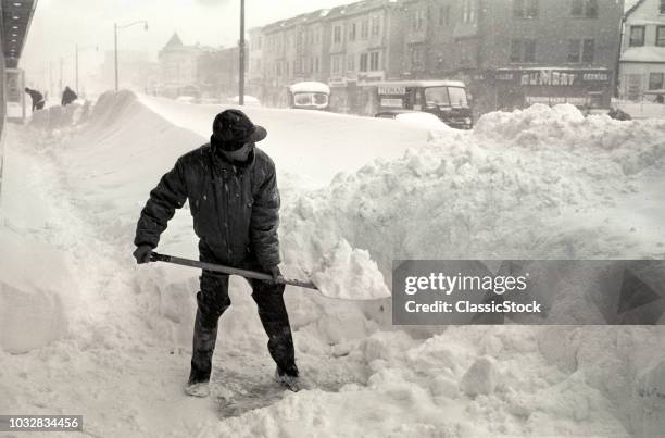 1940s BUNDLED UP ANONYMOUS SILHOUETTED MAN WITH SNOW SHOVEL DIGGING OUT CLEARING CITY SIDE WALK DURING WINTER STORM