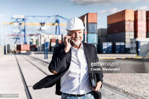 businessman at cargo harbour, wearing safety helmet, using smartphone - friendly match stockfoto's en -beelden