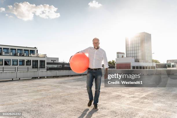 mature man playing with orange fitness ball on rooftop of a high-rise building - german film ball 2018 stock-fotos und bilder