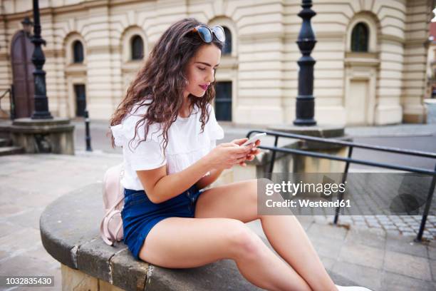 young woman sitting on a wall using cell phone - women in daisy dukes stock pictures, royalty-free photos & images