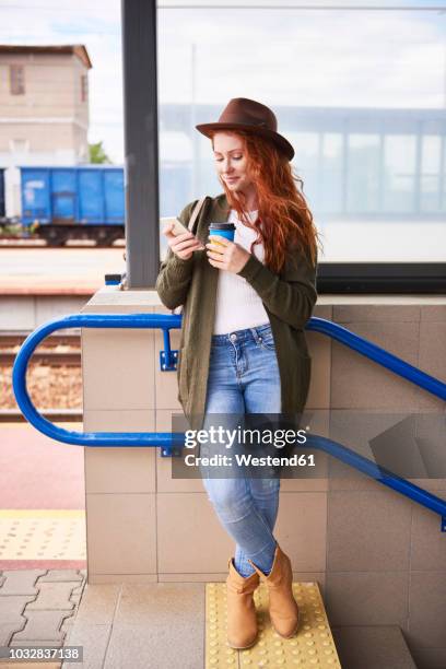 smiling redheaded woman with coffee to go looking at cell phone at platform - human skin cell stock pictures, royalty-free photos & images