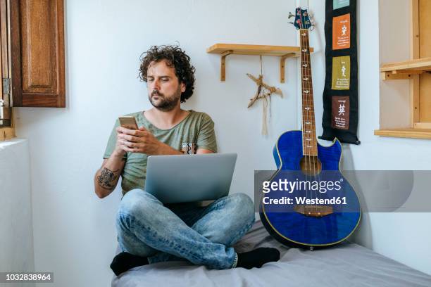 man in his room with mobile phone, laptop and bass guitar - violão acústico - fotografias e filmes do acervo